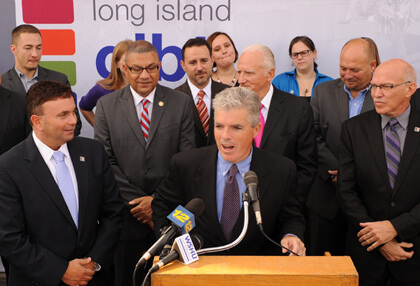 Suffolk County Executive Steve Bellone at the podium during an October 7 ceremony announcing the launch of a LGBT-friendly senior housing development in Bay Shore, with Assemblymember Phil Ramos to the immediate left of the podium and the Long Island GLBT Services Network’s David Kilmnick next to Ramos. | COURTESY: LONG ISLAND GLBT SERVICES NETWORK